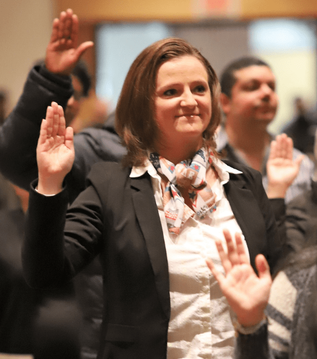 A white woman with shoulder length brown hair raises her right hand during a naturalization ceremony to become a U.S. citizen.