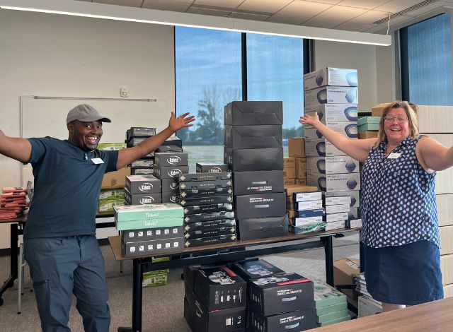 A black man and white woman display boxes of donated kitchen items for refugees.
