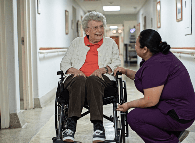 a woman in purple scrubs kneels next to a senior woman in a wheelchair