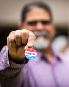 A blurry man with a white beard holds a sticker that says I voted.