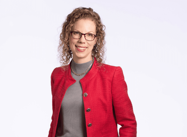 A woman with curly blond hair, glasses and a red business jacket