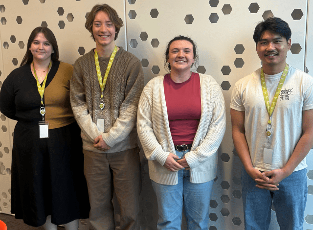 Four college interns stand smiling against a white wall