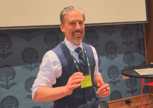 White man with goatee, wearing a blue vest and tie teaching a class
